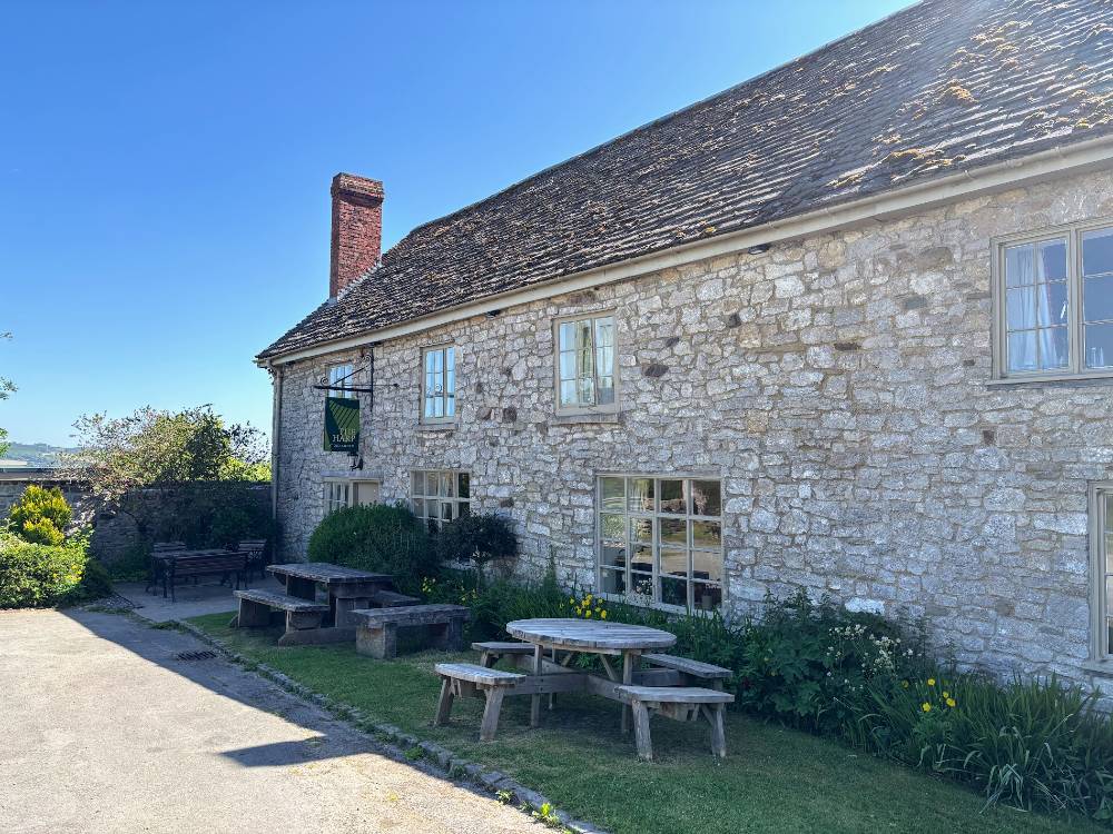 A stone building with a sign 'the harp inn' picnic benches outside. blue sky in the background